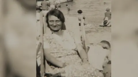 An old sepia-tone photograph of a woman at the seaside. She is wearing a floral dress and sitting in a deck chair. 