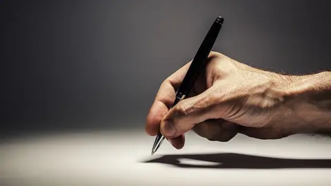 A close-up image of a right hand holding a black pen, poised to write on a blank white surface. The hand is positioned in the center of the image, with the fingers gripping the pen and casting a shadow on the surface below. The background is gradient gray, transitioning from dark at the top to lighter at the bottom