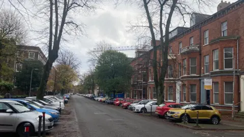 An empty street with cars parked on both sides. On the right side of the street is a row of renovated brick buildings, some of which have scaffolding. On the other side there are larger buildings. There are trees on both sides.  