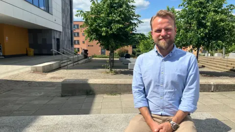 Amy Holmes/BBC A picture of a man who is sat down in the sunshine, with trees and buildings behind him. He is wearing a denim blue shirt and light brown trousers and has short light brown hair and a beard of a similar colour.