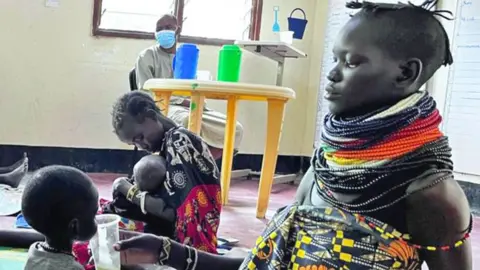 Two women sit on the floor of a ward at Amusait Hospital and feed milk to their young children. One woman has strings of black, white, yellow, orange and red beads around her neck. Both are wearing colourful wraps. A man wearing a blue medical mask sits at a nearby yellow plastic desk, on which there are two jugs and a cup, and looks on.
