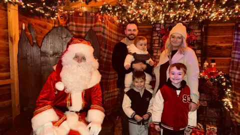 Sarah Wilson A woman and man stand with their three children in a Santa's Grotto, next to a man in a chair dressed as Santa. One of the children is a baby strapped to the father's chest. The other two are young boys. They all smile at the camera.