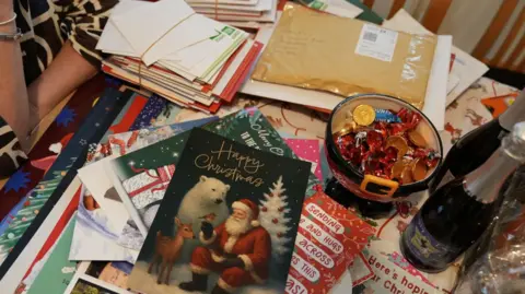 Photograph showing piles of Christmas cards on a kitchen table. In the far left corner there are a stack of Christmas cards tied up with string. A bowl of Christmas chocolates and sweets can be seen in the centre, as well as two bottles of processo. 