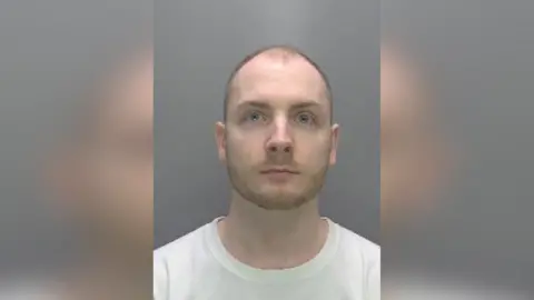 A mugshot of Thurston, who wears a white T-shirt and has a stubbled brown moustache and beard. He stares at the camera.