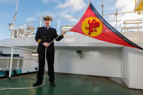 Robert Perry A man in naval uniform raises a red flag with a rampant lion motif on the deck of Glen Sannox