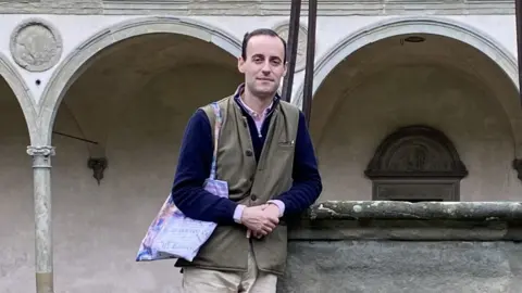 The Victorian Society Connor McNeill is leaning against a stone casket. He has medium length dark receding hair, and is wearing a pink shirt, a blue jersey and a tweed-style long waistcoat in a green colour. He is holding his hands, and has a cloth bag over his right shoulder. There are a series of arches and a monumental plaque behind him, the location of which has not been provided.