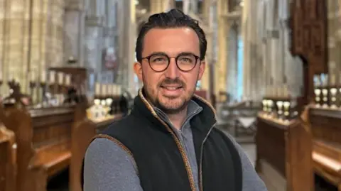 Peterborough Cathedral Jack Pishhorn, a man with brown hair and glasses, is wearing a sleeveless fleece and a grey T-shirt. He is standing in the cathedral and smiling for the camera.