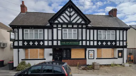 The pub features a mock-Tudor architectural style, with black timber framing against a white plastered exterior and a grey slate roof with two chimneys.