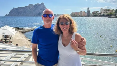 Handout Phil and Anna Brown standing with their arms around each other, smiling at the camera and in front of the sea on a sunny day. Anna is wearing a white dress and sunglasses and Phil is wearing a blue t-shirt and sunglasses. 