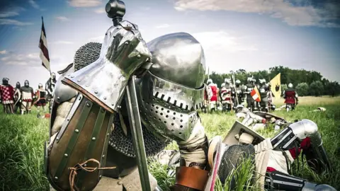 Getty Images A field with a knight in armour kneeling close to the camera, with other knights lying in the grass and other soldiers standing in the background