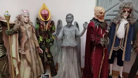 Paul Hayes/BBC An exhibition display of mannequins dressed to appear as various creatures from the BBC science fiction series Doctor Who. The central figure and focus of the photo is a Weeping Angel, a creature designed to look like a stone statue of the type which might be seen in a graveyard. It has its arms raised and spread, and its mouth open to reveal a sharp-toothed scream.