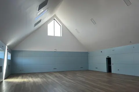 Jack Maclean/BBC An empty sports hall is shown with a vaunted white ceiling and high windows, with blue padded walls and wood-effect vinyl flooring.