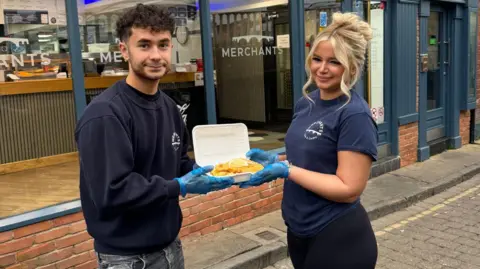 Mitch and Destiny outside Merchants chip shop. They are holding a fish and chip meal and smiling for the camera. 