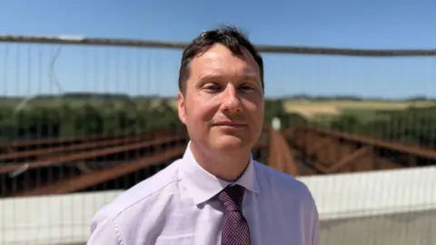 a man with short brown hair wearing a light purple shirt and spotty mauve tie stands in front of metal fencing in front of iron girders forming the base of a bridge