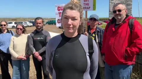 BBC/Jenny Kirk A group of people stand outdoors holding protest placards, a woman in a wetsuit is closest to the camera