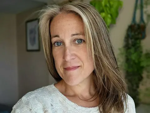 Poppy Whiteside Poppy Whiteside smiling slightly at the camera with her home, with a framed piece of art and plants on the wall, in soft focus in the background