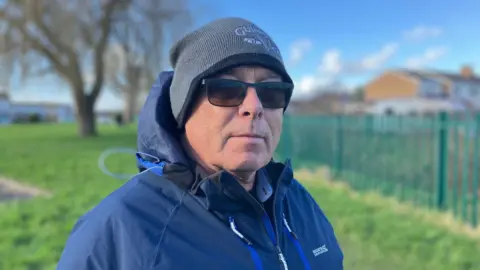 Darren Lynes stands on a footpath in the South Littleton residential estate. He is wearing a grey hat, sunglasses and waterproof coat. Trees and homes are visible in the background