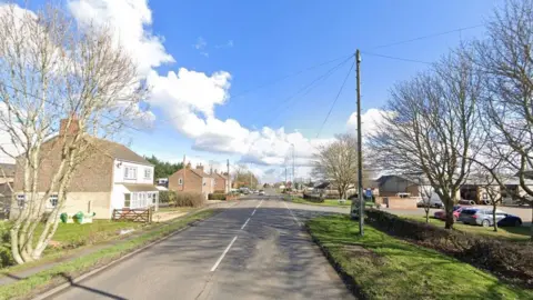 A Google street view of an empty road with a car park on the right and a few detached houses on the left.