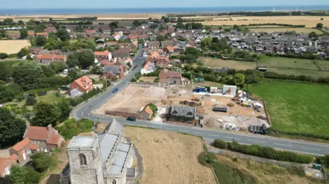 An aerial view of a village with a church in the foreground and a building site behind it with the sea in the distance.