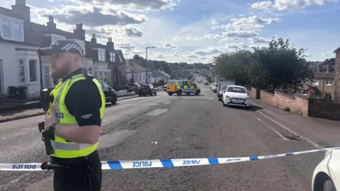 Wide road with a police van half way down between two areas of police tape. A police officer is standing next to tape in the foreground. He has a beard and is wearing a hi-vis jacket and a black and white police cap.  