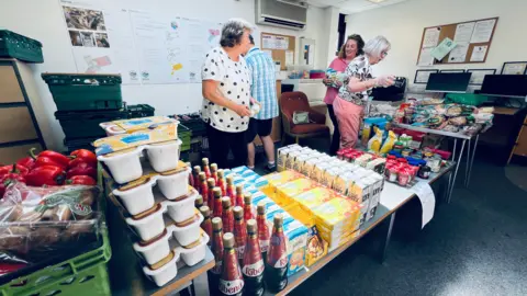 Four volunteers - three women and a man - volunteering at a foodbank. They are organising the food and drink products - which include bottles of Ribena, cartons of oat milk, and snap pots of spaghetti hoops - neatly onto a table.
