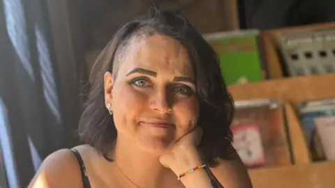 A picture of Beth smiling at the camera. She has black hair and is wearing a bracelet on her wrist. 