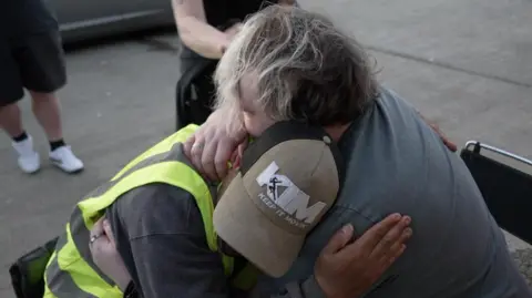 Shaun Whitmore/BBC Mitchell hugs Peter Smorthit following his trek. Mitchell sits on a chair while Peter is in his wheelchair.