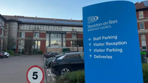 Stockton Council's headquarters Dunedin House. It is a four-storey building mostly made of brown tiles and glass, with solar panels on the roof. A blue sign in the foreground is pointing towards the reception and parking. There are several parked cars and 5mph red and white speed restriction sign.