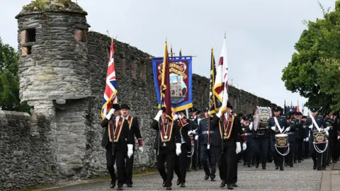 Pacemaker A group of men wearing black suits and black bowler hats marching in a parade. They are wearing maroon sashes an carrying various flags. There is a band behind them and old walls to the left.