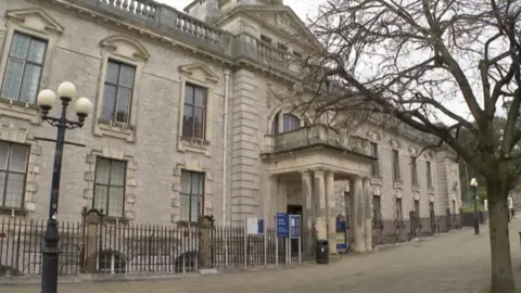A picture outside of the Torquay town hall. It’s a large brick building with a small tree in front of it. It has many windows and gates around it. 
