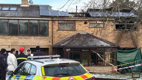 The full house which looks to be semi-detached with another house on the left. The house on the right has a large hole in the roof with beams sticking up. On the ground floor there is a large conservatory. To the right there is a green sheet covering an entrance to the house. There is red and white and yellow and black tape around the garden in front of the house. There is a police car parked in the foreground and a group of five emergency service workers standing behind it. Two of them are in white hazmat suits.