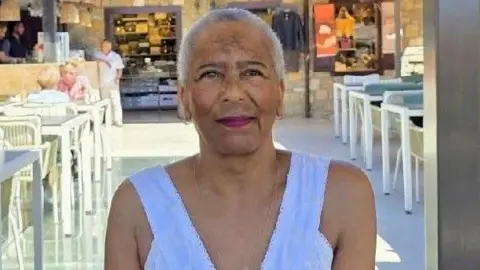 Ruanda Davis A woman with short grey hair is smiling while sitting at an outdoor table. She is wearing a white vest top and some other people can be seen seated at a table a few rows behind her. What looks like a cafe can be seen in the background.