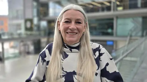 Kay Mason Billig is standing on a balcony in a glass-fronted building. She is wearing a blue and white patterned long-sleeve top.