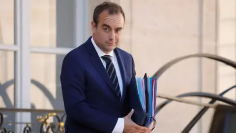 EPA French Prime Minister Sebastien Lecornu leaves the Elysee Palace following the Cabinet meeting in April. He is wearing a blue suit and is carrying a collection of folders. 