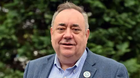 Getty Images Alex Salmond is looking into the camera. He is wearing a light blue checked shirt and slate grey suit jacket with an Alba Party badge pinned to a lapel.