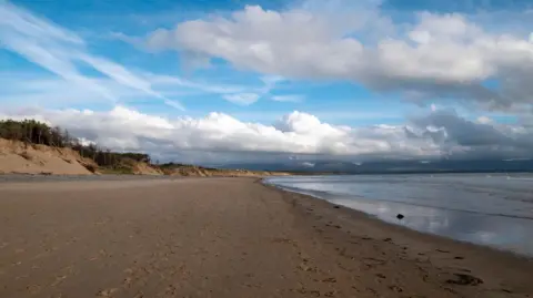 Getty Images The foreground of the image features a wide sandy beach, with footprints scattered across the surface, suggesting recent visitors.
On the left side, there are low sand dunes topped with sparse vegetation and a few trees.
To the right, the calm sea gently laps against the shore with small waves.
The sky is mostly clear blue, with scattered white clouds and some wispy streaks stretching across it.
In the distance, mountains rise up, partially shrouded in clouds, adding depth to the landscape.