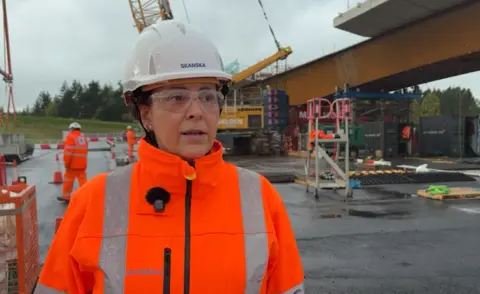 A screenshot of a video interview with Rosario Barcena at the site where the bridge is being built. She is wearing a white hard hat, safety goggles and orange high-viz jacket. She is looking in the distance while being interviewed by someone out of shot. She has a microphone pinned to the side of her jacket. Behind her are heavy plant machinery and workmen in high-viz clothing working around the steel structure of the new bridge.