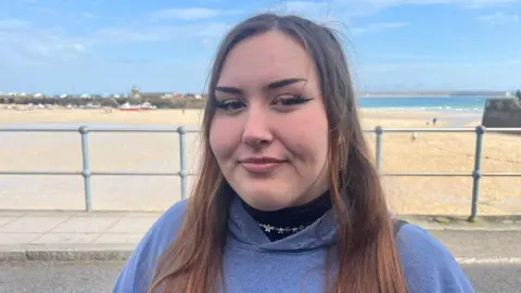 A woman with brown hair, wearing a purple hoody, in front of a seaside backdrop