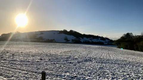 BBC Weather Watchers/DRStevens Sebuah lapangan yang tertutup salju dengan latar belakang bukit di bawah matahari terbit yang cerah.