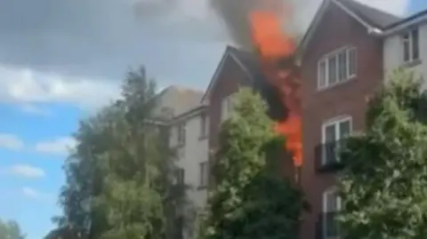 Bright orange flames come out of the side of a block of flats. And smoke can be seen billowing into the blue sky.