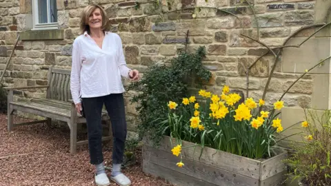 Helen Holmes is a woman in her 60s or 70s, wearing jeans and a white shirt and standing by a trough of daffodils 