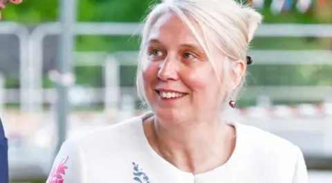 SATH A woman with pale blonde hair and a white top looking upwards slightly