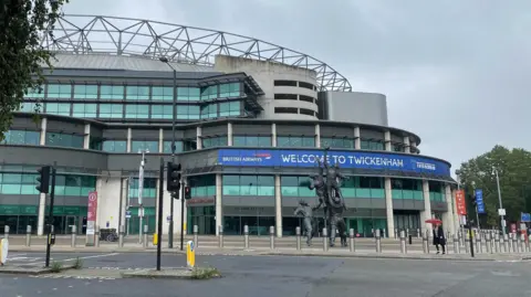 Image shows the front entrance of Twickenham stadium. There is a blue banner that displays welcome to Twickenham and has a British Airways logo on the left. There is a bronze statue of a rugby line-out in front of the stadium. There are traffic lights and a pedestrian crossing visible on the left of the image. 