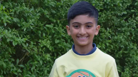 Kayaan has dark hair and eyes and is smiling at the camera. He is wearing a yellow jumper and is standing in front of a hedge.