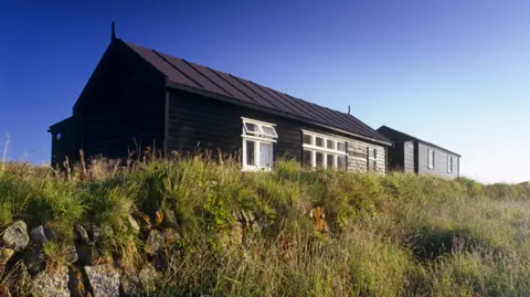 A wide shot of the radio station. It is a brown single-storey hut surrounded by grass.