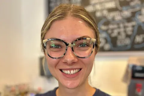 A woman with blonde-hair pulled back and large glasses wearing a dark blue round-necked T-shirt