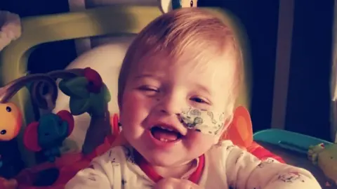 A little boy with blonde hair sat in a high chair. There are colourful toys on his left and he has a feeding tube which is covered by a plaster decorated with dinosaurs.