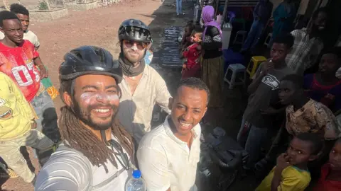 Contributed A man wearing a black cycling helmet takes a selfie with the camera pointed above him. Other people stand around him and also smile at the camera.