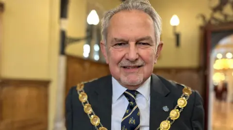 Emma Baugh/BBC Andy Bullen standing inside King's Lynn town hall. He is wearing a gold mayoral chain over a dark suit, white shirt and tie. He has swept back grey hair and a short grey goatee beard. 