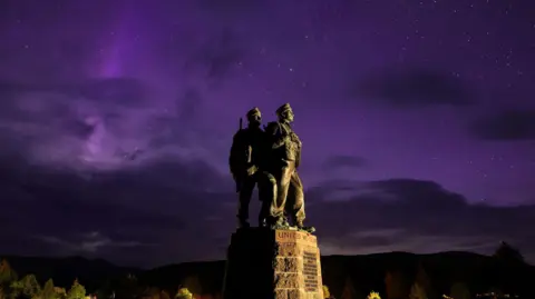 The aurora borealis glows purple behind the Commando Memorial at Spean Bridge. Two of the figures on the memorial are visible. The figures are portrayed wearing military fatigues.